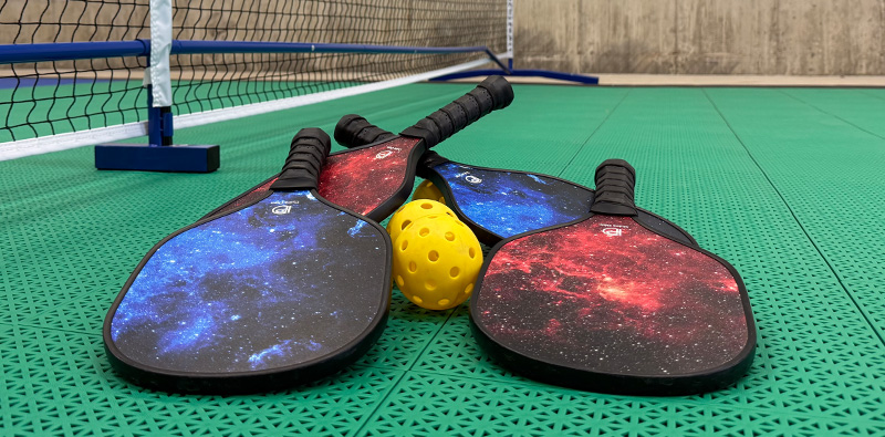Close up of pickleball paddles and whiffle balls under a net.